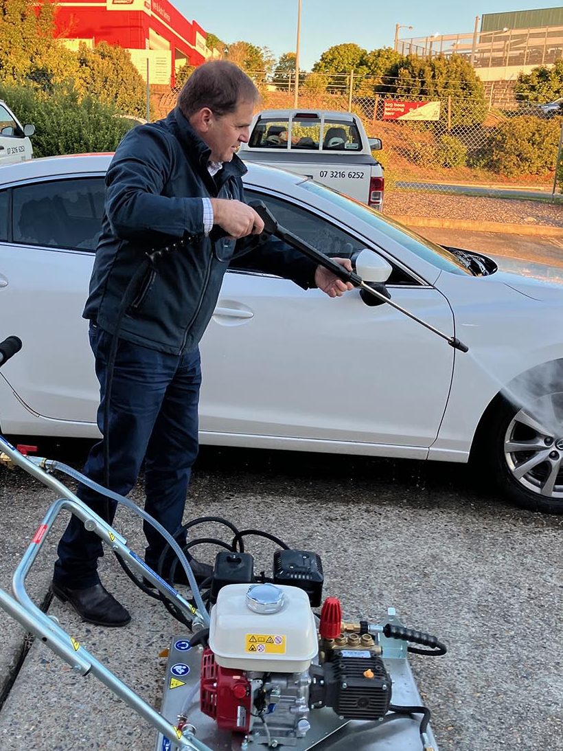 A man using a high-pressure cleaner on a white car in a parking area, with a petrol engine pressure washer visible in the foreground.